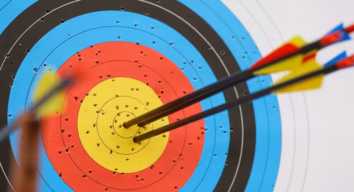 A close-up of a colorful World Archery target face with several arrows clustered in the central gold and red rings.